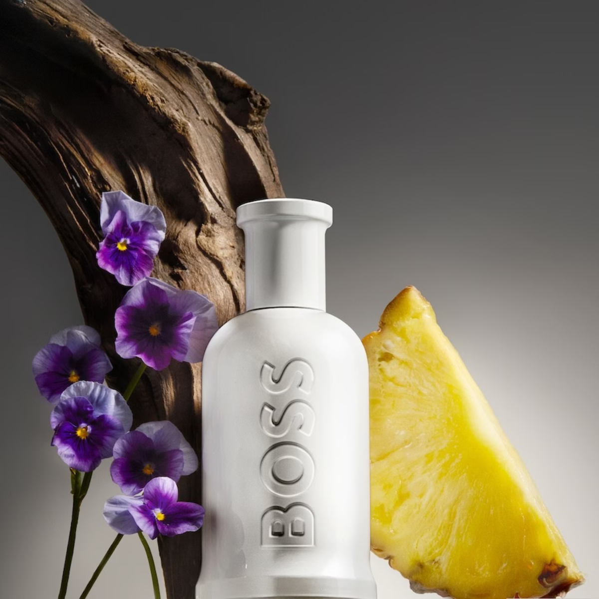 White Hugo Boss bottle with purple flowers and a pineapple slice on a dark background