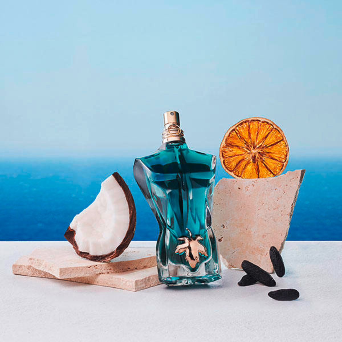 Blue glass bottle with a cork, dried orange slice, coconut shell, and black stones on a blue gradient background.